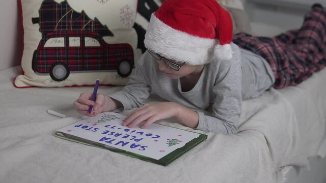 A cute boy for the New Year's red cap writes a letter to Santa Claus asking to stop covid-19 and draws funny pictures on the poster.