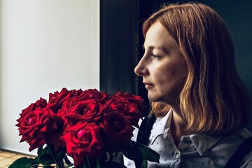 Young woman sits and looks out the window with a bouquet of red roses in her hands