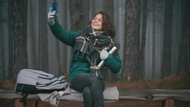 Young cheerful woman tourist taking a selfie on her smartphone with a thermos in hands while sitting on a wooden bench next to her backpack in a misty autumn forest.