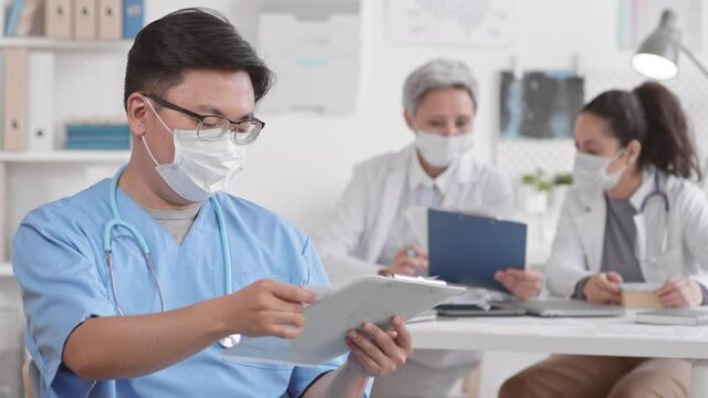 Chest up POV of young male Asian doctor wearing medical mask and blue uniform holding clipboard with papers, leafing through them, looking up on camera, blurred colleagues working on background