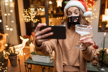 Young woman in facial mask celebrating alone New Year holidays at home, having a video call on the phone with friends. Concept of quarantine and self-isolation during the epidemic on holidays