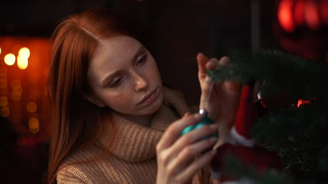 Close-up face of unhappy redhead young woman decorating Christmas tree alone at living room with festive interior. Lonely female preparation her home to holiday celebration during coronavirus pandemic