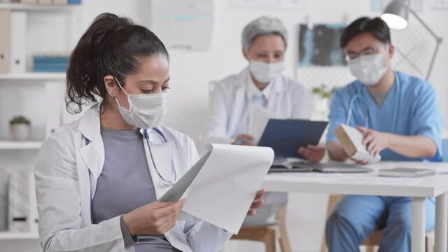 Waist up POV of young female Mixed Race doctor wearing medical mask and white coat holding clipboard with papers, leafing through them, looking up on camera, blurred colleagues working on background