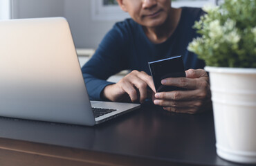 Asian casual man using mobile phone during working on laptop computer on table at home office