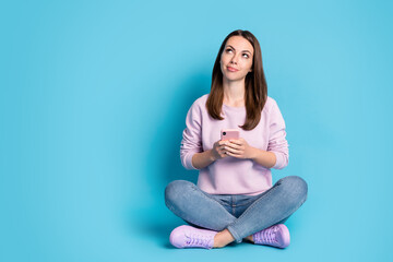 Fototapeta na wymiar Full body size photo of lovely young girl sitting floor crossed legs hold telephone look up empty space smiling wear violet pullover sneakers denim isolated blue color background