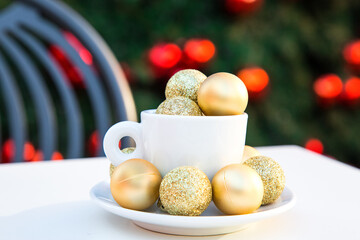 Horizontal picture of coffee cup with golden Christmas balls inside and around. Coffee pair on white table angle, grey chair, new year tree with red toys in background. Winter cafe. Post card picture