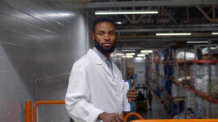Portrait of african engineer in white coat holding clipboard and looking at camera ib warehouse
