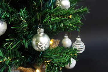 round shiny gray Christmas toy with a snowflake hanging on a branch of an artificial Christmas tree against the background of other toys and glowing garlands on a dark background top view