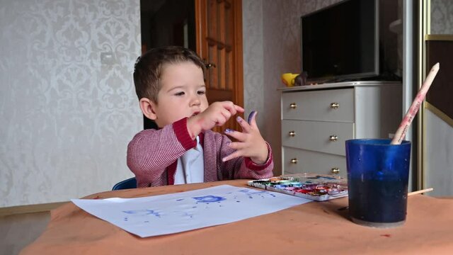 A handsome two-year-old boy sits at the table and counts his fingers on his hand.Children's desk with drawing accessories. Preschool education. 4k