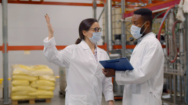 Interracial warehouse inspectors in safety mask and lab coat discussing inventory