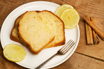Bizcocho en rodajas sobre un plato redondo blanco junto a rodajas de limón, canela en rama, y un tenedor sobre una mesa de madera rústica. Vista de superior