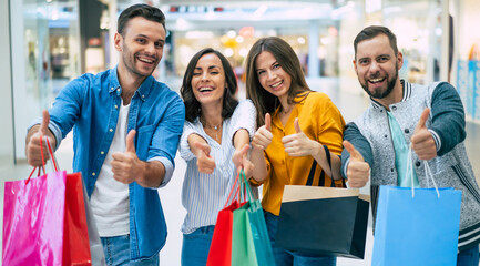 Happy company of smiling excited beautiful stylish friends with colorful paper bags are having fun and showing thumbs up on camera while shopping in the mall.