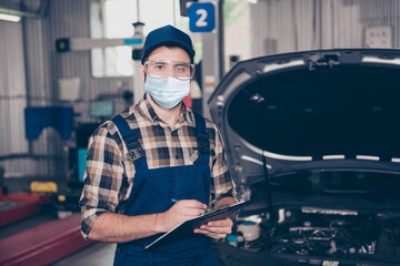 Portrait of skilled guy mechanic writing list car repair maintenance wearing gauze mask goggles in garage