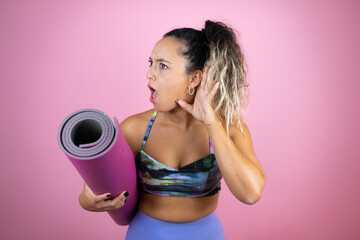 Young beautiful woman wearing sportswear and holding a splinter over isolated pink background surprised with hand over ear listening an hearing to rumor or gossip