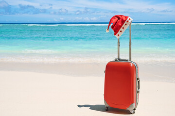 Red Santa Claus Christmas hat on handle of red travel luggage with tropical beach and turquoise sea...