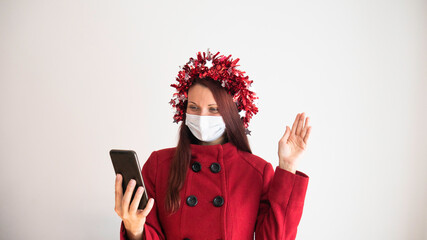Woman in a red coat with a medical mask and a holiday ribbon on her head holding phone and talking online video call during holiday season. Zoom in with family and friends during the flu season 