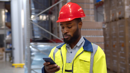 African worker using smartphone standing in large industrial warehouse