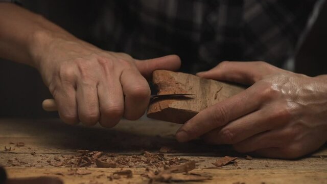 Close up of a Craftsman hands carve with a carving knife a piece of dark wood on carpentry workbench 