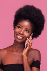 Beautiful African American woman talks on phone or smartphone with gorgeous afro hairstyle standing smiling in black shoulder cut top isolated on pink background. 