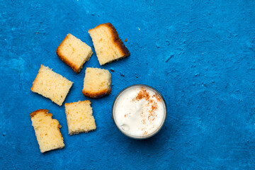 Un vaso de café con leche cn espuma y canela junto a unos trozos de bizcocho sobre un fondo azul clásico y rústico. Vista superior. Copy space