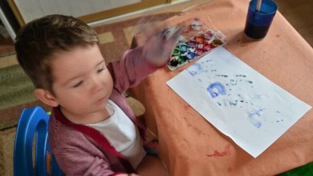 A child looks at the camera and shows his painted hand close-up. Cute little boy draws at home.
