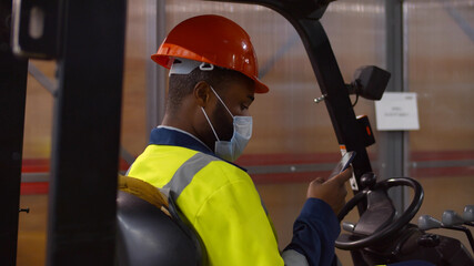African warehouse worker wearing helmet and safety mask sitting in forklift using smartphone © nimito