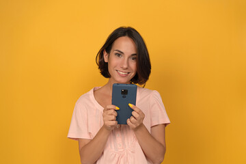 Portrait of beautiful girl holding her smartphone or gadget and cheerful toothy smile on her face while chatting on web or checking social media. Isolated on yellow background. 