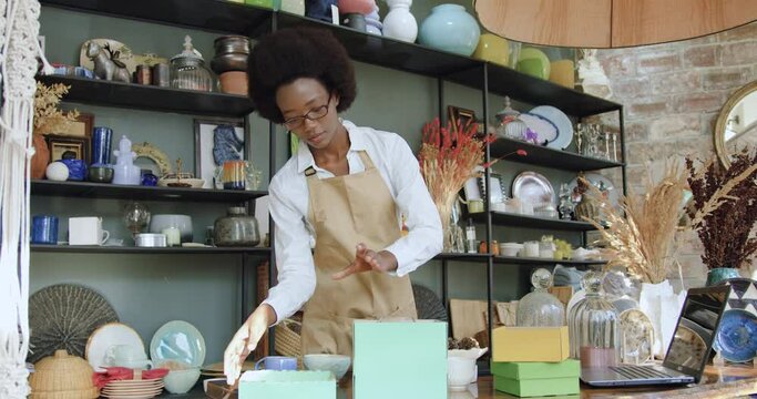 Front view of attractive concentrated confident african american woman which working in beautiful souvenir shop and packes wine glass and cup into gift boxes
