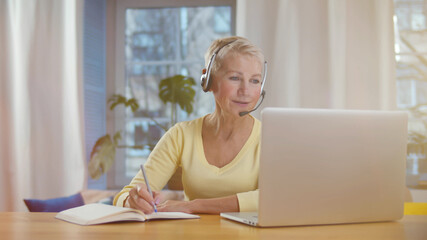 Mature woman with headset and laptop computer having video conference at home office