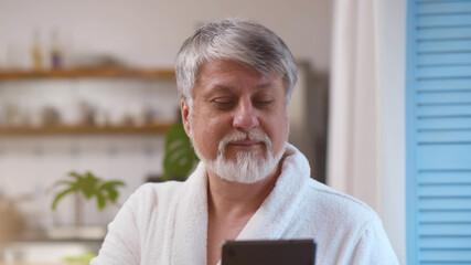 Senior man with a gray beard using digital tablet standing near window at home