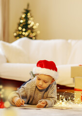 Christmas Child Write Letter to Santa. Kid in Xmas Hat Writing Gift Wish List next to Christmas Tree and home sofa over Background, present boxes