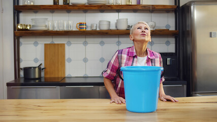 Worried aged woman holding bucket while water droplets leak from ceiling in kitchen