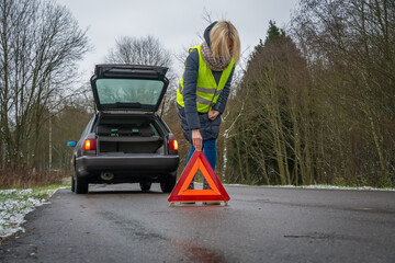 Young blonde woman in a winter down jacket in a yellow vest puts an emergency stop sign on the road an orange triangle near a car with an open trunk
