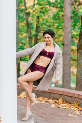 Naklejka premium Young beautiful dark-haired woman with slender figure in lingerie and classic coat in autumn park