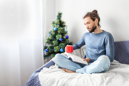 Young bearded man sitting on sofa on blanket reading book and holding cup of coffee near christmas tree at home. Nice to be at home during winter and Christmas holidays