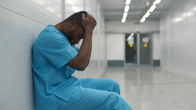 Stressed tired young afro surgeon sitting on floor in hospital corridor after difficult surgery