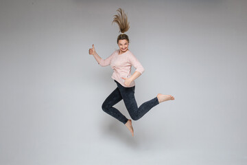 Fototapeta premium Stock photo of cheerful carefree blonde woman jumping in mid-air with thumbs up on white background. Jumping woman smiling at camera holding thumbs up.