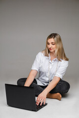 Studio portrait of pretty blonde woman in white shirt, dark jeans and beads on neck typing on laptop sitting with legs crossed on the floor. Studio.