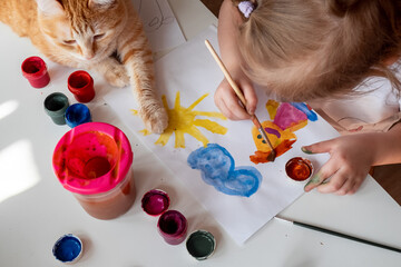 A little girl paints the sun and her mother with watercolors, a ginger cat lies next to the table. Children's creativity. Distance learning, top view