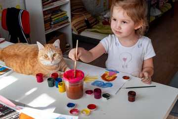 A little girl paints the sun and her mother with watercolors, a ginger cat lies next to the table. Children's creativity. Distance learning,