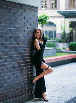 Beautiful young happy glamorous woman in a black evening dress with a gun in her hands. girl in elegant image of spy. High slit on leg. Retro fashion Model posing with pistol on street of modern city.