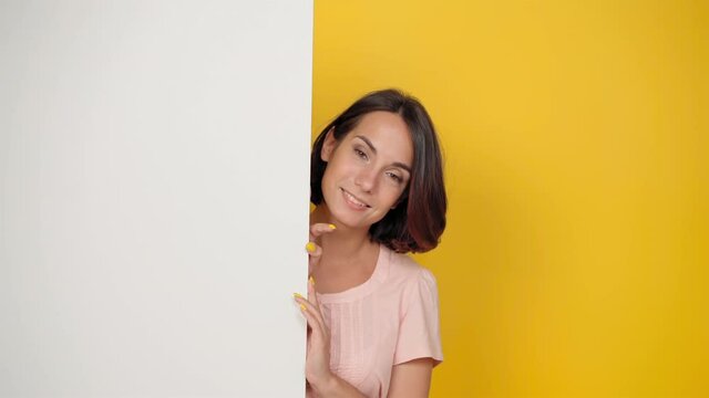 A pretty young girl peeking out of the corner looks at the camera as if watching a family. Smiling young girl in a light pink t-shirt looking out of a corner. Isolated on a yellow background.