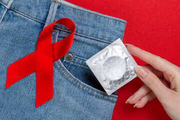A female's hand pulls out a condom from her jeans with a red ribbon. Red background. Flat lay. Close up. The concept of the world day of fight against AIDS and safe sex