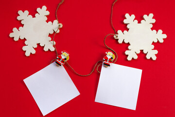 New year's holiday background. White sheets for notes on wooden clothespins with figures of Santa Claus and white empty wooden snowflakes on a red background. Flat lay. Greeting card for Christmas.