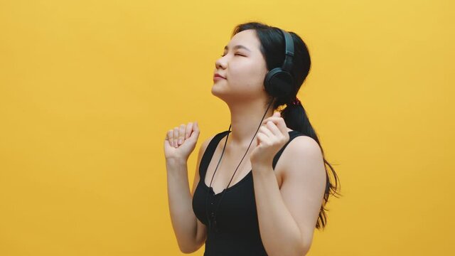 Portrait of happy asian young woman listening to the music on the headphones isolated on yellow background. High quality 4k footage