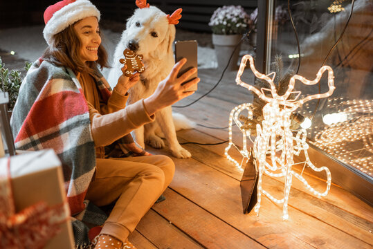 Portrait of a woman in christmas hat and plaid with her cute dog celebrating a New Year holidays at home, feeding dog with gingerbread cookies and making selfie photo