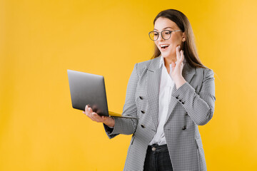 Young woman in jacket and shirt wearing eyeglasses using a laptop on yellow background