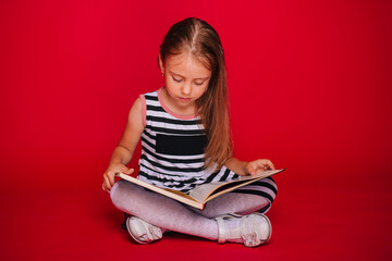 A little girl with blond hair in a striped dress is reading a book. Child on a red background. Learn to read. Preparation for school. Self-isolation lessons