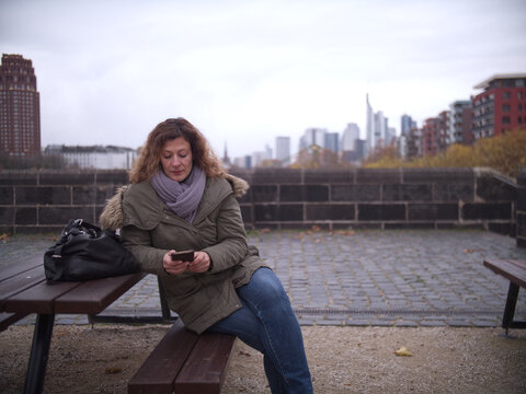 Woman sits on a bench and reads on her mobile phone. Skyline Frankfurt am Main during autumn in the background.