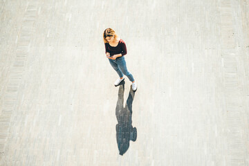 Fototapeta na wymiar young blonde woman in casual clothes walks across empty paved city square looking at modern smartphone screen on sunny summer day upper view